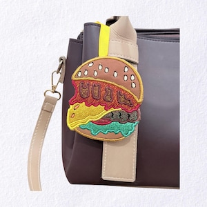 May include: Embroidered burger-shaped bag charm with the word "MUNCH" in brown, yellow, green, and red. The charm is attached to a beige strap and a dark purple handbag. The burger has a brown bun with white sesame seeds.