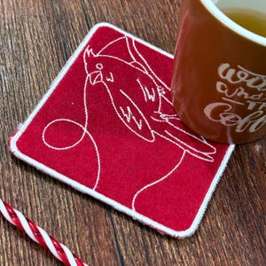 May include: A red square coaster with a white embroidered outline of a cardinal bird. The coaster has a white stitched border. A brown mug with white text is partially visible, resting on the coaster. A red and white striped straw is in the foreground.