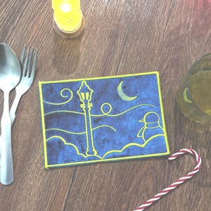 Pode incluir: Uma base azul com um desenho bordado a amarelo com um candeeiro de rua, uma lua e um pinguim. A base é retangular com uma borda amarela. Uma colher, um garfo e uma bengala de doces também são visíveis.