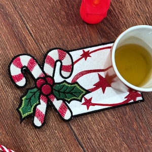 May include: Embroidered Christmas coaster featuring a candy cane, holly, and star design. The coaster is white with red accents and a black border. A white cup with a yellow liquid sits on the coaster.