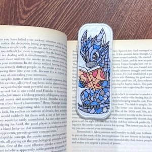Puede incluir: Marcapáginas bordado con un dragón azul leyendo un libro. El dragón tiene detalles intrincados y está sobre un fondo blanco. Un accesorio único para los amantes de los libros.