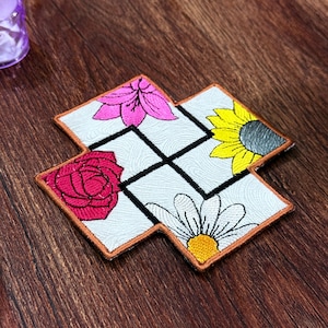 May include: Embroidered coaster with a floral design. The coaster has a white background with a grid pattern and four different flowers: a pink lily, a red rose, a yellow sunflower, and a white daisy. The coaster has a brown border.