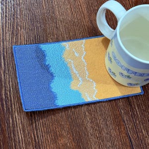 Puede incluir: Un posavasos rectangular bordado con un diseño de escena de playa. El posavasos presenta colores azul, celeste y amarillo, con un contorno bordado blanco. Una taza de cerámica blanca con la palabra "COFFEE" está encima.