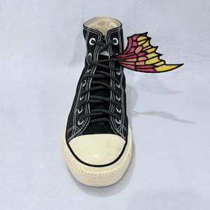 Può includere: Sneaker alta nera con puntale e suola in gomma bianchi. I lacci sono neri. Una toppa ad ala di farfalla colorata nelle tonalità del giallo, arancione e rosso è attaccata ai lacci, aggiungendo un tocco stravagante alla scarpa.