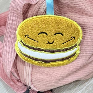 Könnte beinhalten: Ein goldglitzernder Aufnäher in Form eines lächelnden Macarons mit blauem Band. Der Aufnäher hat eine weiße Füllung und braune Akzente. Der Aufnäher ist an einer rosa Cordtasche befestigt.