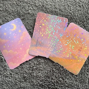 Puede incluir: Tres tarjetas holográficas con ilustraciones de paisajes de ensueño. Una tarjeta presenta un cielo nocturno con una luna creciente y estrellas. Las otras dos tarjetas representan escenas de parque con árboles y bancos, todo con una paleta de colores pastel.