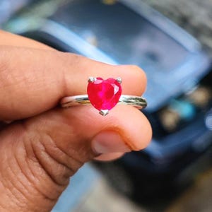 Può includere: Un anello d'argento con una gemma rossa brillante a forma di cuore. La gemma è tagliata per riflettere la luce, creando un effetto scintillante. L'anello ha una semplice fascia in argento lucido.