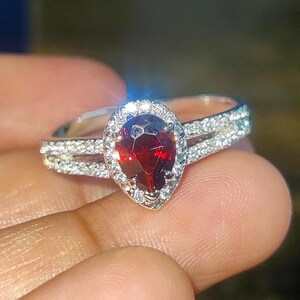 Peut inclure: Une bague en argent ornée d'une pierre précieuse rouge foncé en forme de poire, entourée d'un halo de petites pierres claires. Le corps de la bague est divisé, avec des pierres supplémentaires serties sur les côtés, créant un effet scintillant.