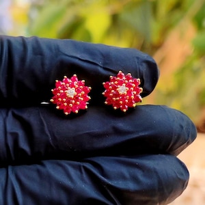 May include: A pair of stud earrings featuring a cluster of vibrant red gemstones arranged in a circular floral pattern, with a single clear white gemstone set at the centre of each piece, mounted on a gold-toned metal base.