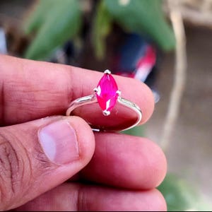 May include: A silver ring with a marquise-cut, bright pink gemstone. The gemstone is held in place by four prongs, and the band is polished silver. The ring is shown against a blurred background of green leaves.