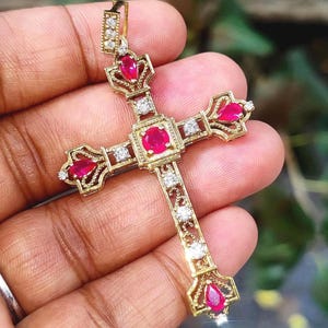 Peut inclure: Un pendentif en forme de croix de couleur or, orné de pierres précieuses rouges et de petits diamants blancs. La croix présente un design détaillé avec des bords ornés et une boucle en haut pour une chaîne. Les pierres précieuses sont serties au centre et aux extrémités.