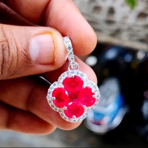 May include: A silver pendant with a four-leaf clover design. The pendant features four bright red gemstones in the centre, surrounded by a halo of smaller, clear gemstones. The bail is also encrusted with clear gemstones.