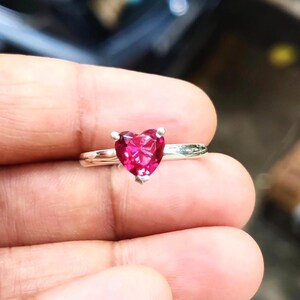 Può includere: Un anello d'argento con una gemma rossa a forma di cuore. La gemma è il punto focale, incastonata su una fascia sottile. L'anello è tenuto in una mano, mostrando il suo design e il suo colore.