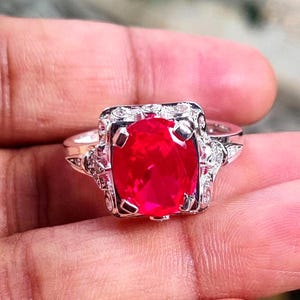 May include: A silver ring with a large, vivid red gemstone. The gemstone is set in a square frame with detailed embellishments and smaller accent stones. The ring's band is silver.