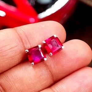 May include: A pair of square-cut, vibrant red gemstone stud earrings with silver settings. The gemstones have a deep, rich colour and are set in a simple, elegant design. The earrings are displayed on a hand.