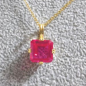 May include: A gold-colored necklace with a square, bright pink gemstone pendant. The pendant is set in a gold frame and hangs from a delicate gold chain. The gemstone appears to be approximately 0.75 inches square.