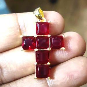 Può includere: Un ciondolo a forma di croce color oro, con pietre preziose rosse scure a taglio quadrato. Le pietre sono incastonate in metallo color oro, creando un contrasto sorprendente. Il ciondolo ha un anello nella parte superiore per una catena.