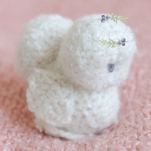 Puede incluir: Un juguete de pollito blanco de crochet con un ojo gris. El pollito está sentado sobre una superficie rosa y peluda.