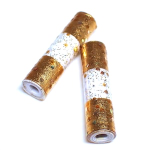 May include: Two decorative rolls of gold and white patterned paper. The gold sections are covered in glitter, and the white sections have small, scattered glitter accents. The rolls are cylindrical and appear to be made of paper.