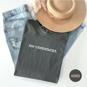 May include: A casual fashion flat lay featuring a gray t-shirt with the text "no comment." The shirt is paired with light blue jeans, a tan hat, and a silver necklace. A small, round sticker with the word "PEPPER" is in the bottom right corner.