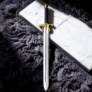 May include: A decorative sword with a silver blade and a gold-colored hilt. The handle is wrapped in black material. The sword is displayed on a white marble surface and a dark gray, fluffy rug.