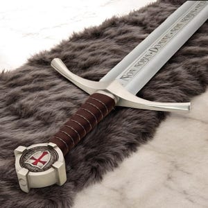 Pode incluir: Uma espada prateada com uma empunhadura envolta em couro castanho e um emblema em forma de cruz. A lâmina tem a inscrição "NON NOBIS DOMINE NON NOBIS SED". A espada está sobre uma superfície de pele sintética cinzenta.