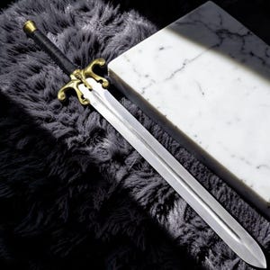 May include: A long, silver sword with a gold-colored hilt and a black handle. The sword is resting on a dark gray, textured surface next to a white marble block. The sword's blade is sharp and pointed.