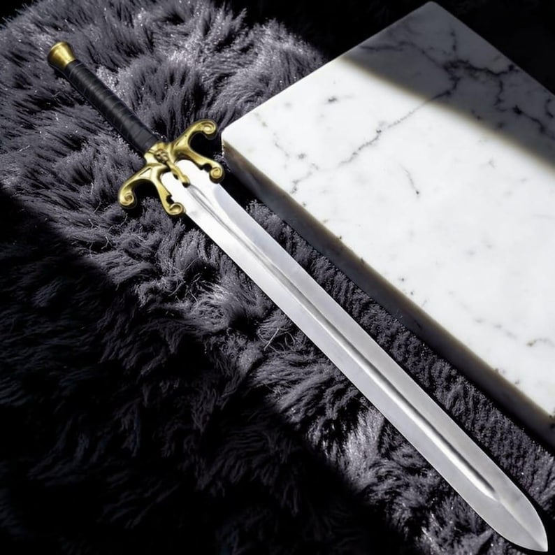 May include: A long, silver sword with a gold-colored hilt and a black handle. The sword is resting on a dark gray, textured surface next to a white marble block. The sword's blade is sharp and pointed.