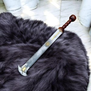 K&ouml;nnte beinhalten: Ein r&ouml;misches Gladius-Schwert mit Holzgriff und Stahlklinge. Der Griff ist dunkelbraun und die Klinge hat einen goldenen Akzent. Auf der Klinge befindet sich ein goldenes Spinnennetz-Design. Das Schwert liegt auf einem dunkelgrauen, pelzigen Teppich.