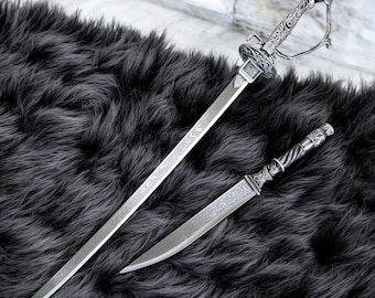 May include: Two ornate silver swords with detailed engravings rest on a dark gray, textured surface. The larger sword has a curved handguard and a decorative pommel. The smaller sword has a twisted handle and a straight blade. The blades are sharp and pointed.