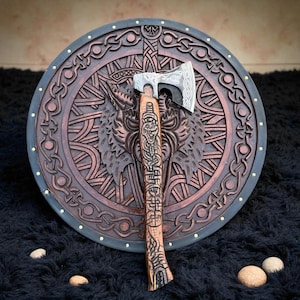 Könnte beinhalten: Eine Axt im Wikinger-Stil mit detailliertem Holzgriff und silberner Klinge liegt vor einem runden, aufwendig geschnitzten Holzschild. Der Schild zeigt ein Wolfsmotiv und keltische Knotenmuster mit dunklem Rand und goldenen Akzenten.