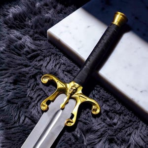 May include: A decorative sword with a gold-colored hilt featuring a skull design and a black leather-wrapped handle. The blade is silver, and the sword rests on a dark, textured surface.