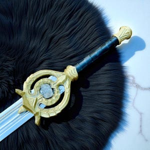 Puede incluir: Una espada decorativa con una empuñadura dorada y una hoja azul grisáceo. La empuñadura presenta un diseño ornamentado con una piedra central. El mango está envuelto en material azul oscuro. La espada se muestra sobre una superficie negra texturizada.