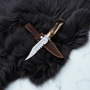 May include: A handcrafted hunting knife with a serrated blade and a deer antler handle. The knife is in a brown leather sheath, resting on a dark, textured surface. The blade has etched details.