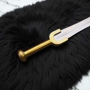May include: A decorative sword with a gold-coloured handle and a silver blade, resting on a black, fluffy surface. The handle has a rounded end and a curved guard. The blade is straight and has a sharp edge.