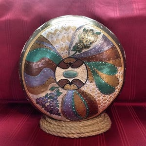 May include: A round, painted gourd with a decorative design featuring swirls, flowers, and geometric patterns in shades of brown, gold, green, purple, and blue. The gourd is displayed on a rope stand.