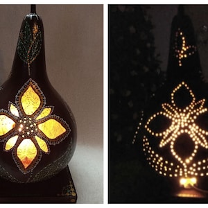 May include: A decorative gourd lamp with a dark brown exterior and a floral design. The lamp has a square base and a long, teardrop-shaped body. The floral design is illuminated from within, creating a warm glow.