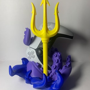Könnte beinhalten: Ein gelber Dreizack wird von einer lila-blauen, oktopusartigen Basis getragen. Hinter dem Dreizack befindet sich ein weiß-schwarzer Gamecontroller. Der Dreizack hat eine leuchtend gelbe Farbe.