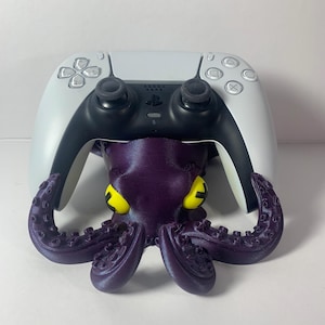May include: A purple octopus-shaped controller stand with yellow eyes, holding a white and black game controller. The stand is designed to cradle the controller, providing a unique and decorative storage solution.