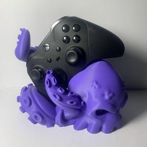 May include: A black video game controller rests on a purple octopus-shaped stand. The controller has multiple buttons and a textured grip. The stand features detailed tentacles and a stylized octopus head, providing a unique gaming accessory.