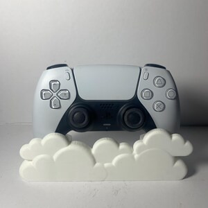 May include: A white and black PlayStation 5 controller with a white cloud-shaped decorative item in front. The controller has black joysticks and buttons with the PlayStation logo.