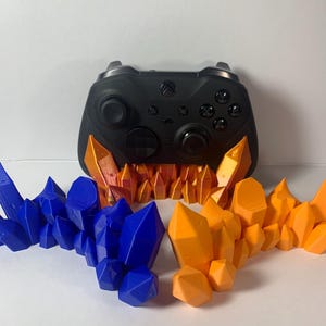 May include: A black Xbox controller rests on a decorative stand featuring blue and orange crystal-like formations. The stand is designed to cradle the controller, with the crystals adding a unique aesthetic. The crystals are arranged in a symmetrical pattern.