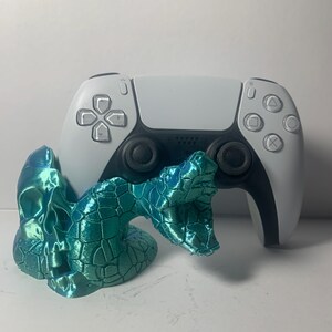 Może przedstawiać: Turkusowa, iryzująca figurka węża umieszczona przed biało-czarnym kontrolerem PlayStation 5. Wąż ma teksturowany, łuskowaty wygląd. Kontroler ma czarne joysticki i przyciski.
