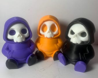 May include: Three seated skull figurines wearing hoodies in purple, orange, and black. Each has a white skull face and black shoes. The purple and black figurines have purple feet, while the orange figurine has black feet.