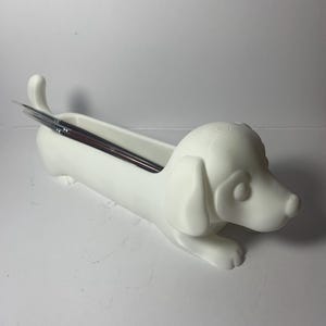 Puede incluir: Un organizador de escritorio blanco con forma de perro salchicha. El organizador tiene un cuerpo largo y rectangular, una cola curvada y una cabeza de perro con orejas caídas. Varios pinceles están dentro.
