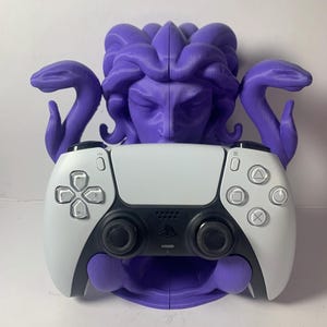 Op de afbeelding: Een paarse Medusa-thema controllerhouder met een wit en zwarte gamecontroller. De houder heeft een gebeeldhouwd gezicht met slangenhaar en armen. De controller heeft knoppen en joysticks.