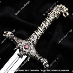 May include: A sword with a metallic silver finish featuring an ornate hilt. The pommel is shaped like a lion head, and the crossguard displays intricate scrollwork with a central square red gemstone. The blade is smooth, polished, and reflective.