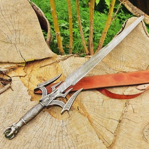 May include: A detailed, ornate sword with a patterned blade and decorative hilt. The handle features a dragon's head design. The sword rests on a wooden surface with a brown leather sheath visible. The blade measures approximately 30 inches.