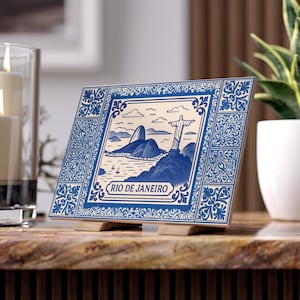 May include: A square ceramic tile featuring a blue and white illustration of Rio de Janeiro. The image includes the Christ the Redeemer statue, mountains, and the text "RIO DE JANEIRO". The tile is displayed on a wooden stand.