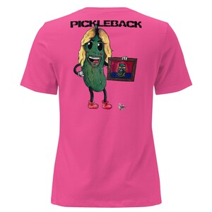 May include: A vibrant pink t-shirt with a cartoon pickle character with blonde hair, holding a framed picture of a jar. The text "PICKLEBACK" is printed in black across the top. The pickle is wearing red shoes.